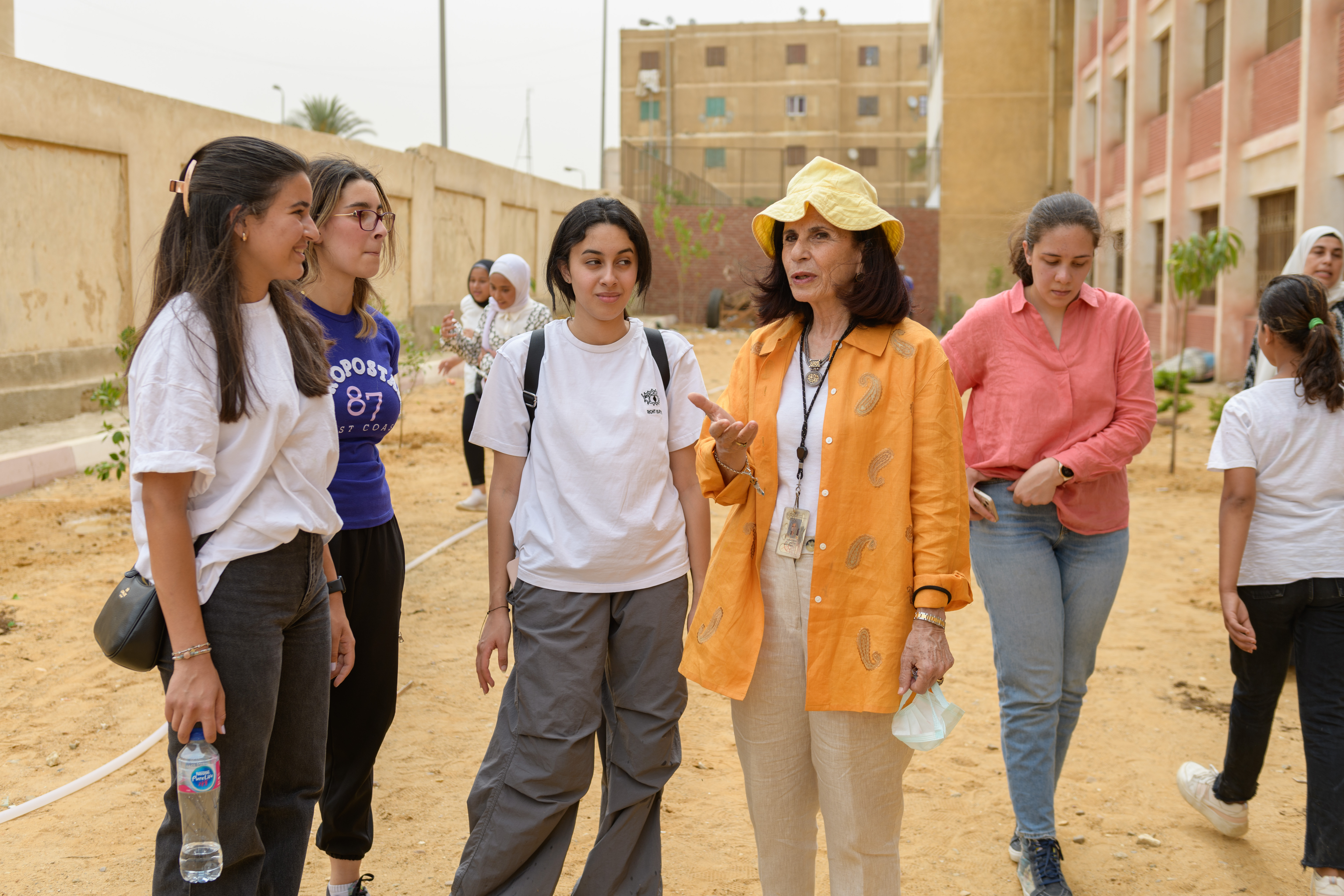 a woman in an orange shirt speaks with students in the yard of a school