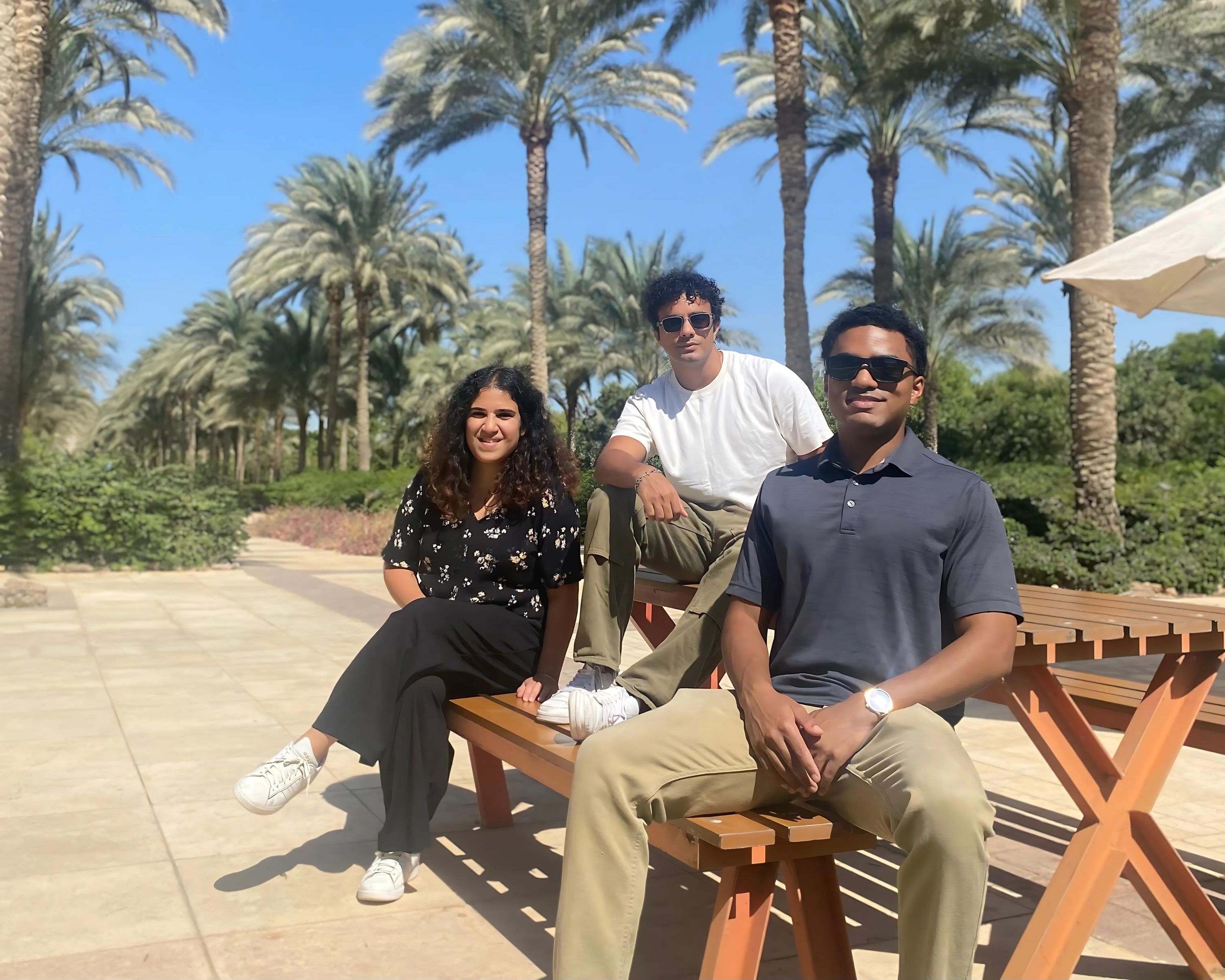 Two men and one woman sit on a picnic table in the AUC garden, palm trees in the background