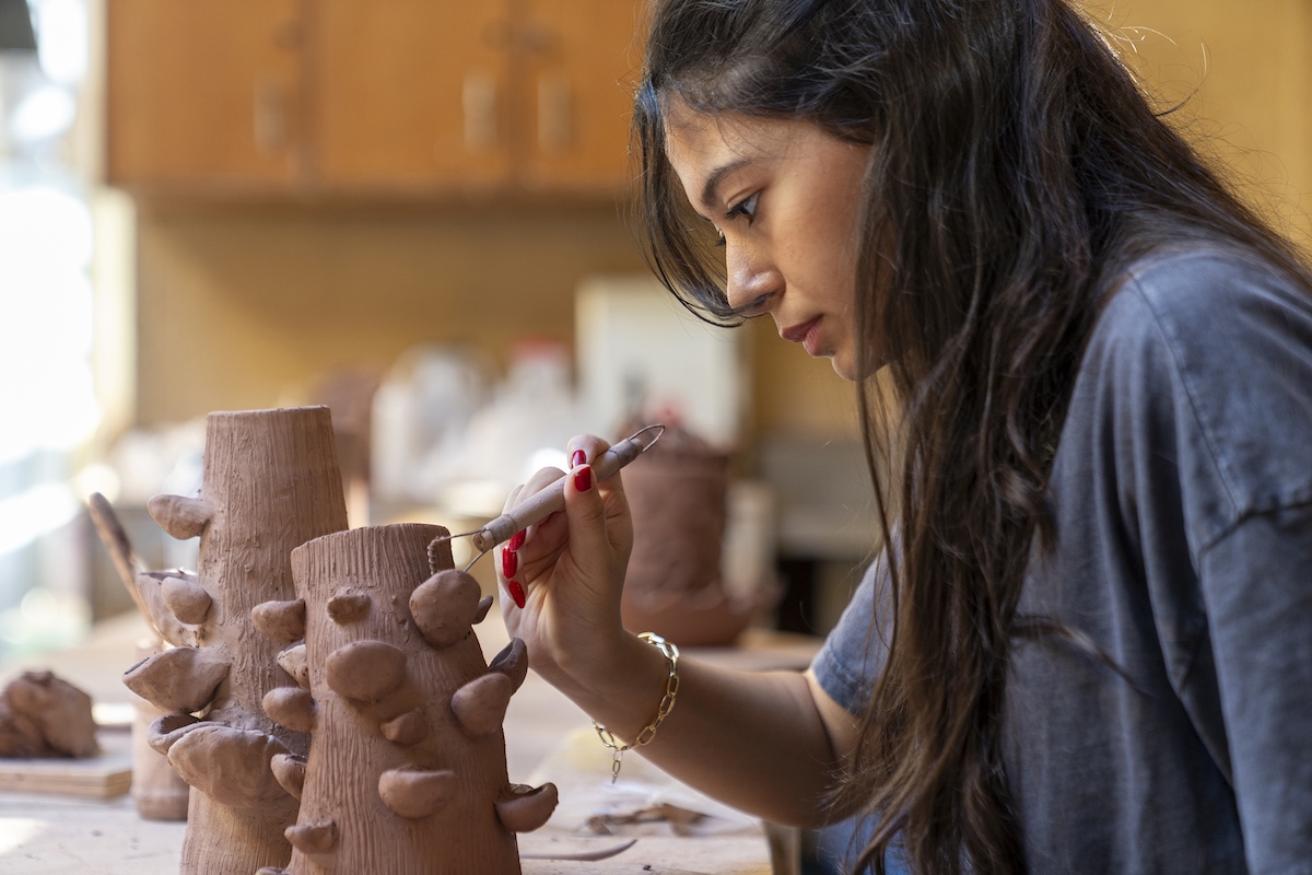 A student carefully shapes and details a clay sculpture in an art studio.