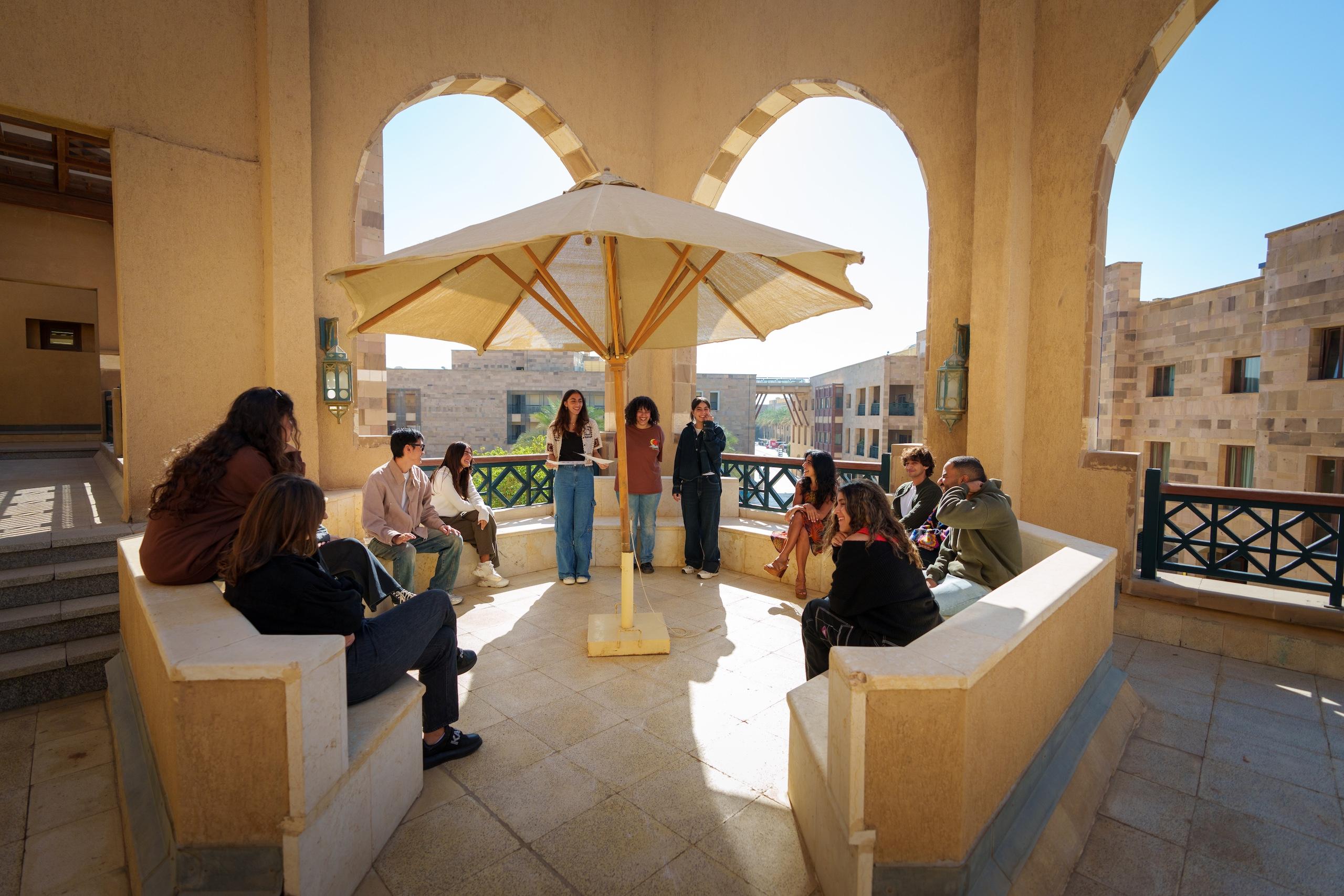 Males and females are standing under a big umbrella in a semi-open space