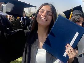 A female is wearing her gown and holding her cap and certificate. She is smiling 