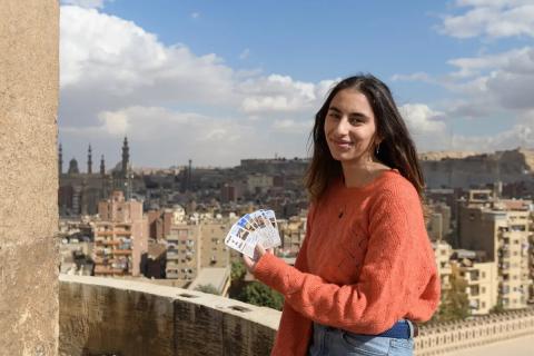 A girl is smiling, she is holding cards with pictures on them. There are buildings and mosques behind her