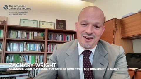 A mans is talking into a camera and he is smiling. He is wearing a suit. There are bookshelves behind him