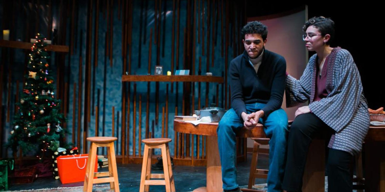 a scene from a play featuring a male and female seated next to each other with the woman's hand on the man's shoulders and a christmas tree in the background