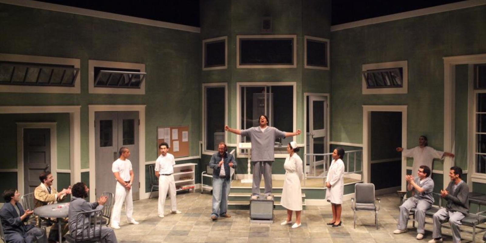 A scene from a play feautring a group of people standing a semi circle and the man in the center is standing on a higher step with his hands opened