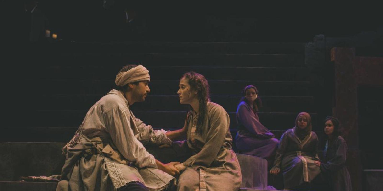 A scene from a theater production featuring a male and female wearing vintage cloths seated opposite to each other on stage