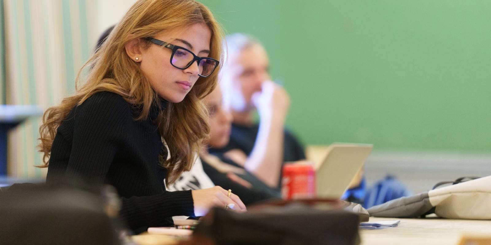 A student wearing glasses concentrates while working on a laptop in a classroom.