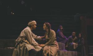 A scene from a theater production featuring a male and female wearing vintage cloths seated opposite to each other on stage