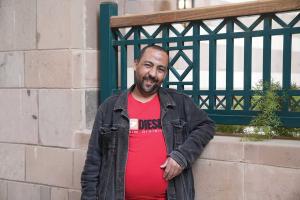 Mohamed Salah, known as Tata, stands smiling outdoors on AUC&rsquo;s campus, leaning against a low wall, wearing a red T-shirt and dark jacket.
