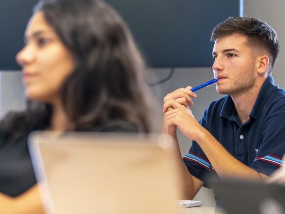 A student listens thoughtfully during a class discussion while holding a pen.